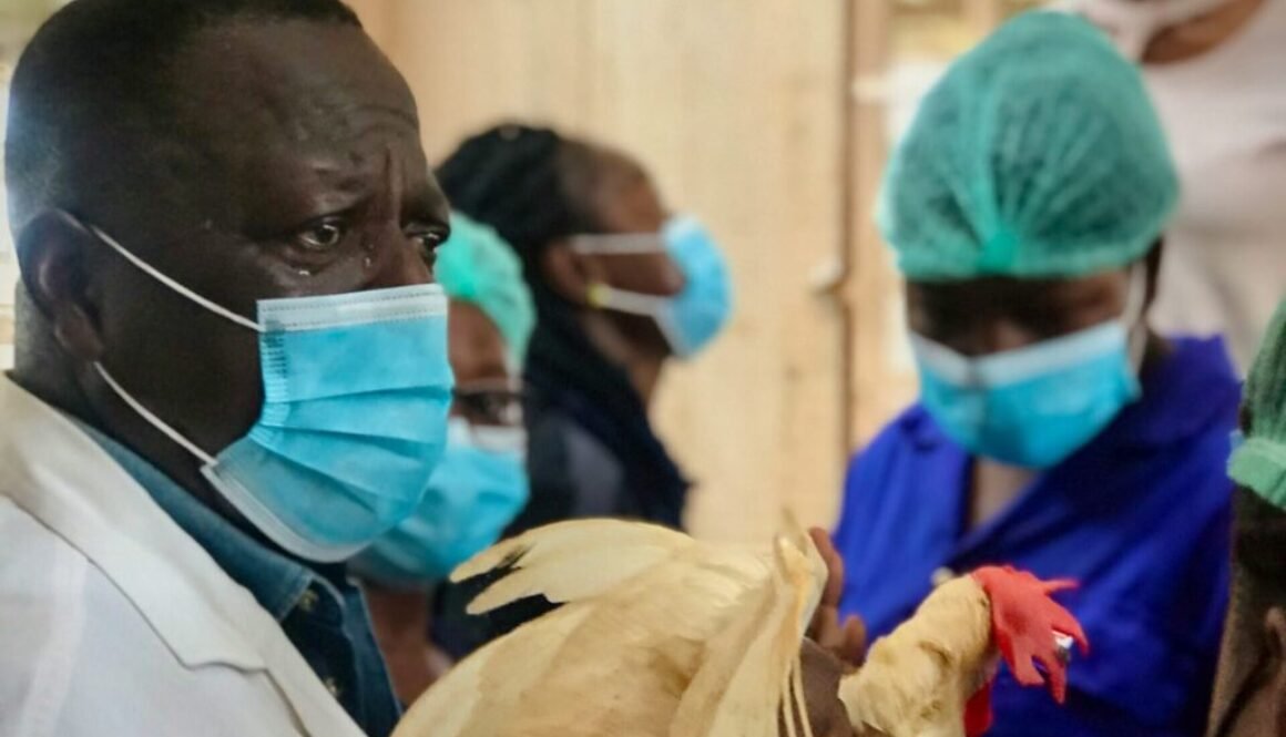 A man in a medical mask holds a chicken while masked workers look on in the background